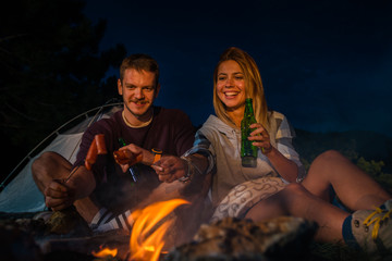 Young couple baking sausages on the campfire and drinking beer in the forest hill in the dusk