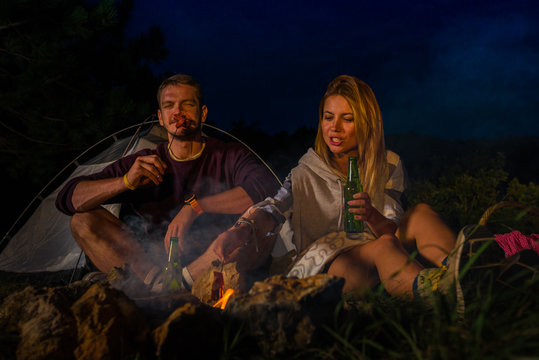 Young couple baking sausages on the campfire and drinking beer in the forest hill in the dusk