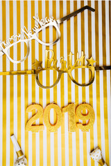 New Years Eve celebration background , 2019 number made with golden glitter canles, flatlay over a striped golden white board, luxury  holiday concept, with funny parts glasses.