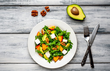 Arugula salad, persimmon, walnuts, avocados, feta cheese on a white plate on a wooden table