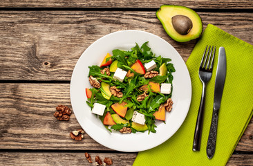 Arugula salad, persimmon, walnuts, avocados, feta cheese on a white plate on a wooden table