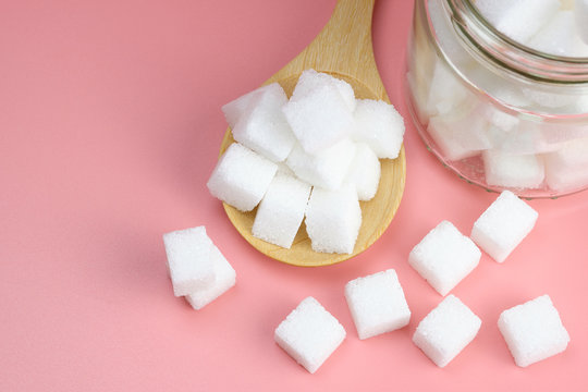 Cube granulated sugar in a glass jar and  on wooden spoon for adding sweetness. some parts was poured on a pink background. - image