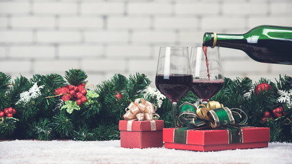 Red wine is pouring from bottle in to glass decorating with red gift boxes and Christamas ornaments. Copy space at top area with blurred white brick wall
