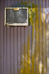 An old box air-conditioning unit hangs from the side of a corrugated iron wall.