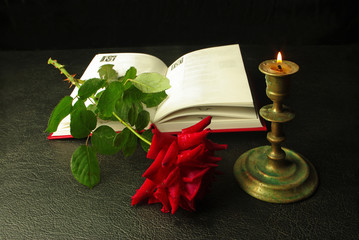 Red rose, an open book and a burning candle. On a black background. 