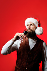 Thoughtful Santa man drink glass of red wine. Businessman in Santa hat holds glass of wine. Bearded man tasting red wine. Handsome rich man with mustache&beard holds glass of red wine. New year party.