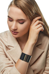 A portrait of a Caucasian girl with long light brown hair, wearing a woolen suit jacket and grossy bangle bracelet. The woman is touching her hair while posing against white background, looking down.