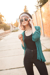 Fototapeta premium Young woman with sunglasses and hat is walking along the street at sunset