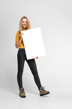 Positive Laughing Woman Wearing Black Jeans And Yellow T-shirt With Blondie Hair, Toothy Smile Is Holding White Big Mockup Poster Isolated On White Background