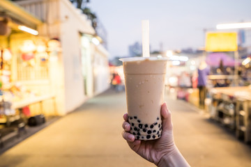 A young woman is holding a plastic cup of bubble milk tea with a straw at a night market in Taiwan,...