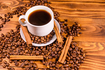 Cup of hot coffee, cinnamon sticks and scattered coffee beans on wooden table