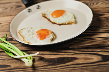 Two fried eggs in metal beige pan and green fresh onion on old wooden brown table. Copy space