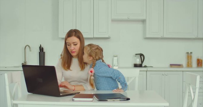 Affectionate caring single mother working laptop, embracing and kissing her cute little daughter with love and happiness in domestic kitchen. Loving freelance mum always has time for her child.