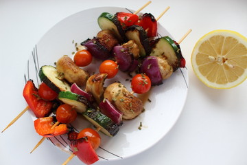 Grilled zucchini on skewers with tomatoes, onion, peppers,  and mushrooms with lemon on a white plate and light background. Vegan food. Top view.