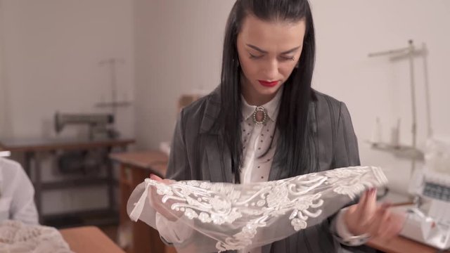 A female designer holding a cutout-veiling sleeve with beaded lace and checking it. She smiles and says something. A tailor is sitting to her left and sewing beads down the veiling cloth.