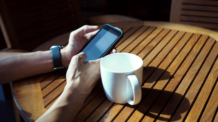 Closeup of Asian male hands playing smartphone and drinking coffee at an outdoor table with morning sunlight.