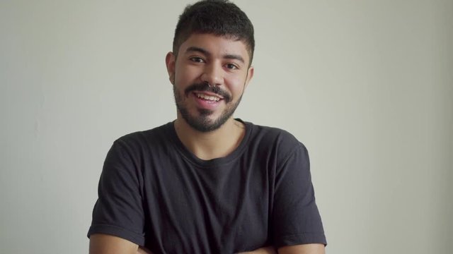 Handsome bearded man with piercing in nose smiling at camera. Happy young man in black t-shirt crossing arms and looking at camera on grey wall background. Happiness concept