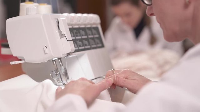 A close up side view of a seamstress hemming white fabric on the sewing machine. She finishes stitching, fastens off the thread and cuts it with scissors