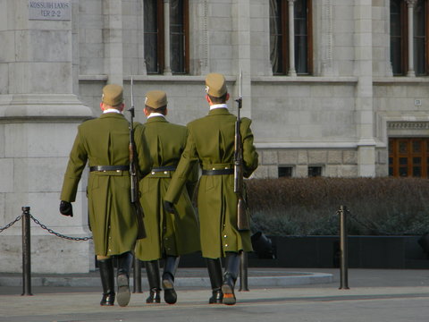 Three members of the Hungarian Honor Guard marching towards the Hungarian Parliament after the changing of the guard near the Hungarian flag on Kossuth Lajos ter, with tourists in the background