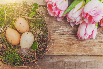 Obraz na płótnie Canvas Spring greeting card. Easter eggs in nest with moss and pink fresh tulip flowers bouquet on rustic shabby wooden background. Easter concept. Flat lay top view copy space. Spring flowers tulips
