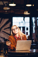Work and travel, study and vacation freelance concept. Vertical of petty blond woman in sweater and eyeglasses using smart phone laptop at evening in loft interior bar, pub cafe. Start up concept.