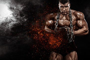 Brutal strong muscular bodybuilder athletic man pumping up muscles with chains on black background....