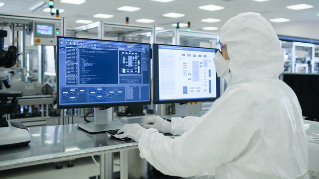 Shot of a Scientists in Sterile Suits Working with Computers, Analyzing Data form Modern Industrial Machinery in the Laboratory. Product Manufacturing Process: Semiconductors, Biotechnology.
