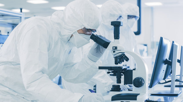In Laboratory Scientists in Protective Clothes Doing Research, Using Microscope and Entering Data into Personal Computer. Modern Manufactory Producing Semiconductors and Pharmaceutical Items.
