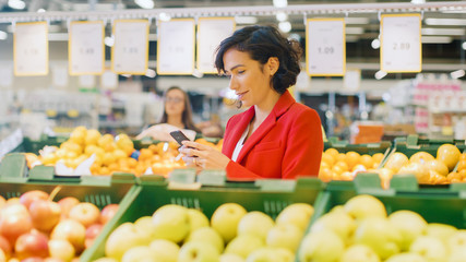 At the Supermarket: Woman Using Smartphone, Chooses Products In the Fresh Produce Aisle. Woman Immersed in Internet Surfing on Her Mobile Phone In the Background Fruits and Organic Vegetables.