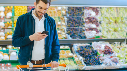 At the Supermarket: Handsome Man Uses Smartphone while Standing in the Fresh Produce Section of the Store. Man Immersed in Internet Surfing on His Mobile Phone. Colorful Fruits and Organic Vegetables.