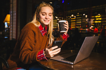 Blond millennial girl using laptop and smartphone in cafe, outdoor portrait, hipster style, cellular, internet, text message, business lady, make call, work. Freelancer doing her business project.