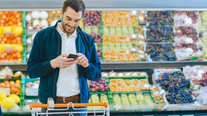 At the Supermarket: Man Uses Smartphone while Standing in the Fresh Produce Section of the Store. Man Immersed in Internet Surfing on His Mobile Phone In the Background Colorful Fruits and Vegetables.