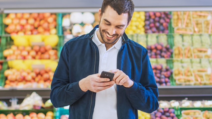 At the Supermarket: Man Uses Smartphone while Standing in the Fresh Produce Section of the Store. Man Immersed in Internet Surfing on His Mobile Phone In the Background Fruits and Organic Vegetables.