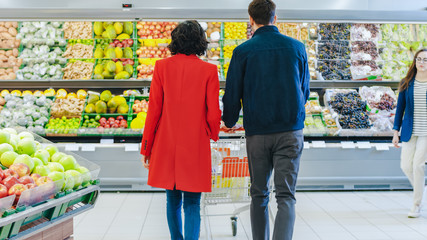 At the Supermarket: Happy Young Couple Chooses Organic Fruits and Berries in the Fresh Produce Section of the Store. Boyfriend Pushes Shopping Cart while Girlfriend Picks up Fruits.