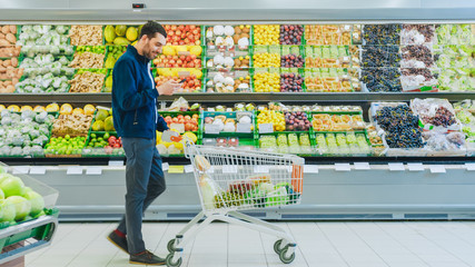 At the Supermarket: Handsome Man with Smartphone, Pushes Shopping Cart, Walks Past Fresh Produce...