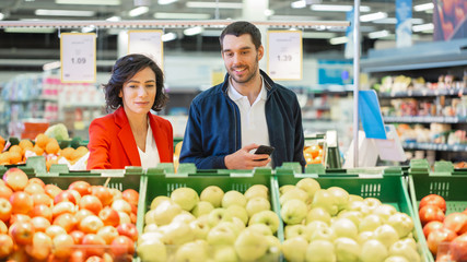 At the Supermarket: Happy Young Couple Chooses Organic Fruit in the Fresh Produce Section of the Store. Husband Uses Smartphone, Wife Picks Up Fruit. 
