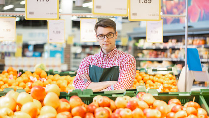 At the Supermarket: Portrait Of the Handsome Stock Clerk Wearing Apron, Arranging Organic Fruits and Vegetables, He Smiles and Crosses Arms. Friendly, Efficient Worker at the Store.