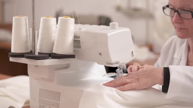 A dressmaker is hemming white fabric on the overlock sewing machine to make a wedding dress. A woman is adjusting the cloth, and saying something in the end