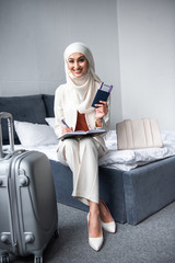 beautiful young muslim woman holding passport with boarding pass and smiling at camera at home