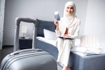 close-up view of suitcase and smiling muslim woman holding passport with boarding pass at home
