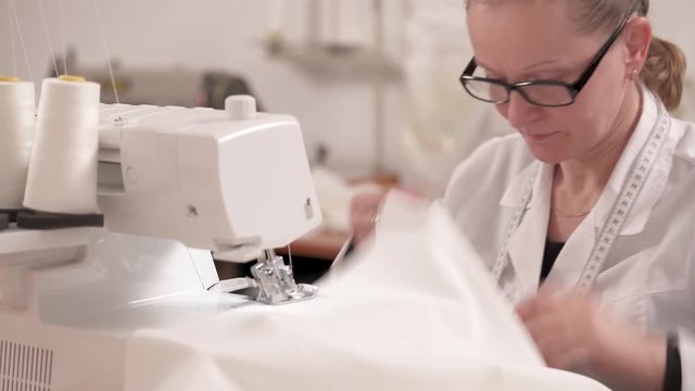 A woman in glasses and white gown is sitting and is working on the overlock sewing machine to hem white fabric. Work in the atelier to make a wedding dress