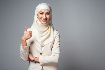 young muslim woman smiling at camera and showing thumb up isolated on grey