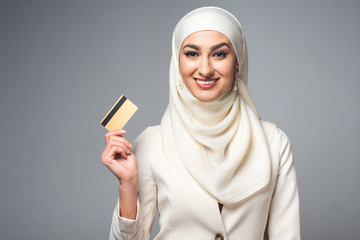 beautiful muslim woman holding credit card and smiling at camera isolated on grey