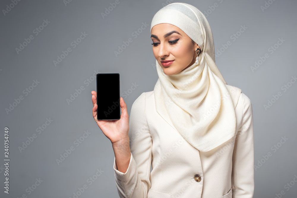 Wall mural beautiful smiling young muslim woman holding smartphone with blank screen isolated on grey