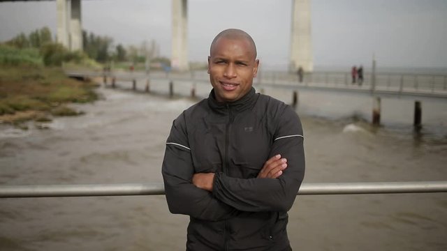 Smiling hairless sportsman with crossed arms standing on pier. African american man wearing sportswear looking at camera. Healthy lifestyle concept