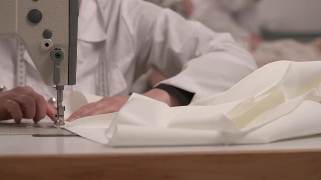 A close-up of woman's hands finishing to hem white fabric on the overlock sewing machine to make a wedding dress. She trims the edges with scissors and cuts the thread off in the end