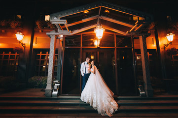 Tender and lovers newlyweds hugging against the background of the night city and light bulbs with bokeh. Stylish groom and beautiful bride with a long dress on the stairs.