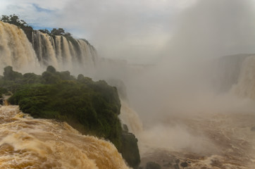 Iguazu falls on the brazilian side with some green bushes, water mist and a cloudy sky