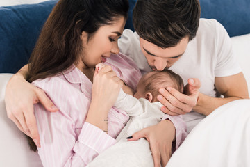 young mother breastfeeding little baby with husband near by on bed at home