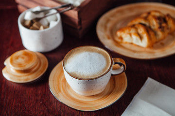 A cup of morning cappuccino and freshly baked pecans on a plate of warm flowers on a dark wood table